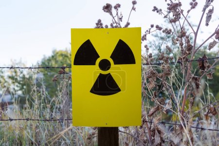 Foto de Ionizing radiation hazard sign in the form of black trefoil on yellow sheet nailed to the post on a background of the fence of rusty barb wire and dry burdock and other grass - Imagen libre de derechos