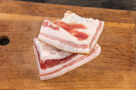 Two pieces of the salted pork fatback on skin with meat layers lie on wooden cutting board, close-up