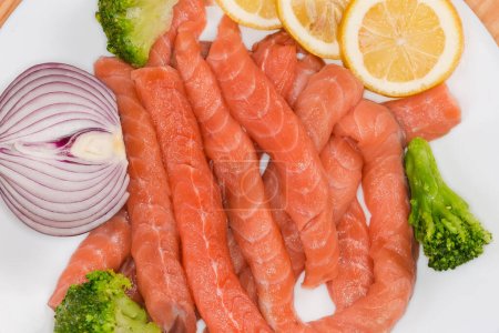 Small long slices of the raw salmon with vegetables pieces and lemon slices for preparation of fish casserole on a white dish, top view close-up