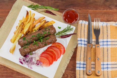 Photo for Serving of the baked lula kebab with French fries, chopped vegetables and sauce on the white square dish on an old rustic table, top view - Royalty Free Image