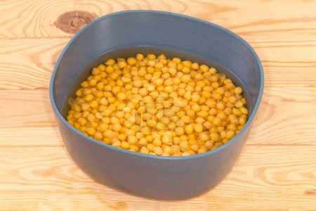 Téléchargez les photos : Pois chiches jaunes entiers non cuits trempés dans l'eau froide dans le bol bleu-gris avant de se préparer à une cuisson plus rapide sur la table rustique - en image libre de droit