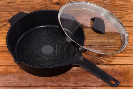 Empty deep frying pan made of cast aluminum with titanium non-stick coating with partly removed lid of glass and stainless steel on the rustic table