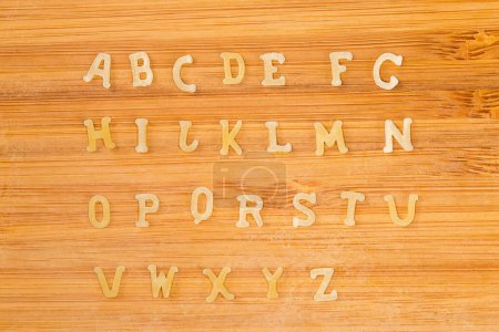 Téléchargez les photos : Pâtes crues en forme de lettres majuscules de l'alphabet anglais placées en rangées dans l'ordre alphabétique sur la planche à découper en bambou, vue de dessus gros plan - en image libre de droit
