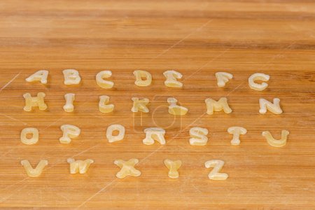Téléchargez les photos : Pâtes crues en forme de lettres majuscules de l'alphabet anglais placées en rangées dans l'ordre alphabétique sur la planche à découper en bambou, vue de côté gros plan - en image libre de droit
