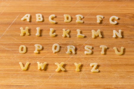 Téléchargez les photos : Pâtes crues en forme de lettres majuscules de l'alphabet anglais placées en rangées dans l'ordre alphabétique sur la planche à découper en bambou, gros plan - en image libre de droit