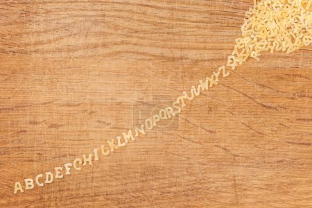 Foto de Pasta cruda en forma de letras mayúsculas del alfabeto inglés colocada en línea en el orden alfabético estirada a partir de un montón en la vieja tabla de cortar, vista superior - Imagen libre de derechos