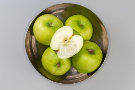 Photo for Half of the green apple lies on the same whole apples in the stainless steel kitchen bowl on a gray background, top view - Royalty Free Image