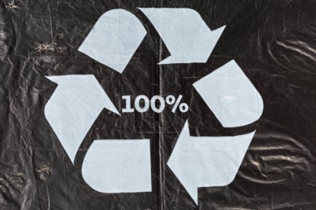 White universal recycling symbol and inscription 100% on the black slightly wrinkled food plastic bag made from the recycled plastic, close-up
