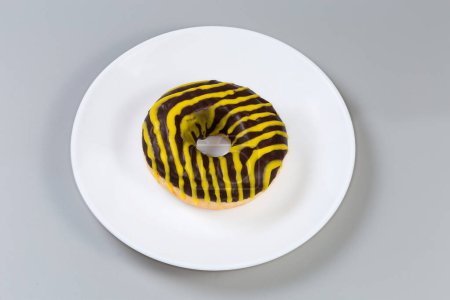 American-style ring shaped doughnut with striped chocolate and yellow glaze on a white dish on a gray background