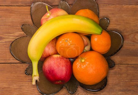 Photo for Different fresh washed apples, banana and tangerines in a vintage glass vase for fruits on the old rustic table, top view - Royalty Free Image