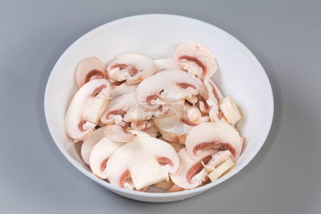 Fresh raw button mushrooms chopped into thin slices in white bowl on a gray background