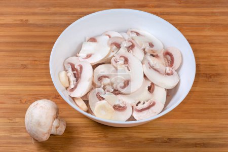 Fresh raw button mushrooms chopped into thin slices in white bowl on a wooden surface