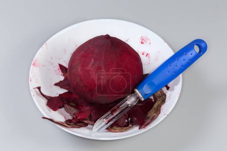 Remolacha roja redonda cruda entera pelada de la piel y peladora de verduras entre los peelings en un tazón sobre un fondo gris