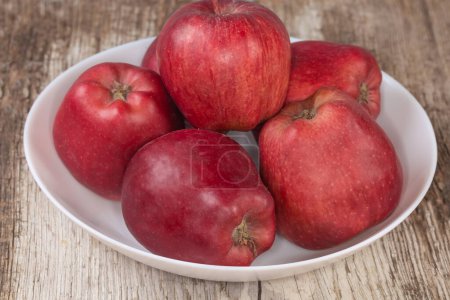 Photo for Whole dark red apples on the white dish on an old cracked wooden surface, side view close-up in selective focus - Royalty Free Image