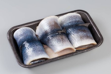 Pickled herring fillets on skin rolled into a cylindrical shape on the black foam food tray on a gray background