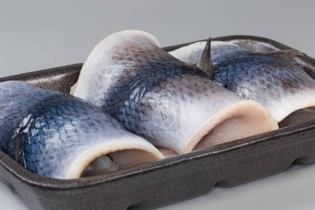 Pickled herring fillets on skin rolled into a cylindrical shape on the black foam food tray on a gray background, side view close-up
