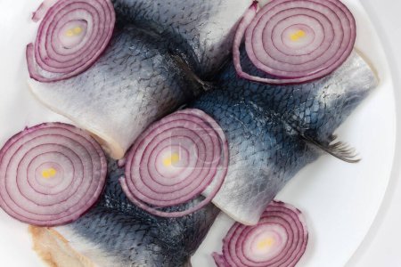 Pickled herring fillets on skin with fresh red onion sliced into rings on the white dish, top view close-up