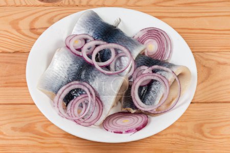 Pickled herring fillets on skin with fresh red onion sliced into rings on white dish on a rustic table