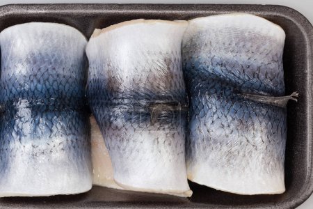 Pickled herring fillets on skin rolled into a cylindrical shape on the black foam food tray, top view close-up
