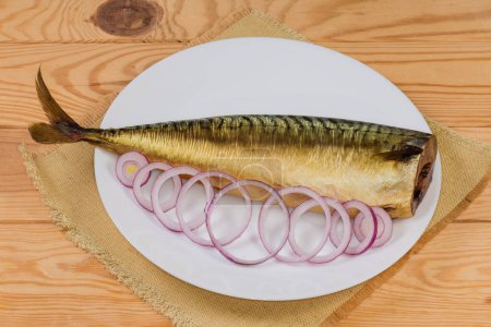 Whole cold-smoked Atlantic mackerel without head with fresh red onion sliced into rings on a white dish on a rustic table