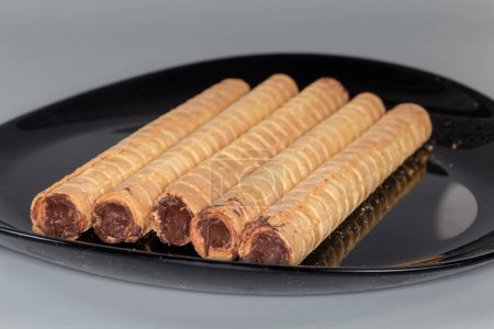 Fragment of the black dish with wafer tubes with filling of chocolate cream on a gray background, side view close-up in selective focus