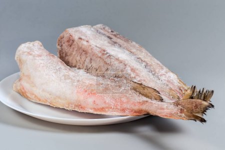 Frozen carcasses of the red cod without heads, covered with hoarfrost on white dish on a gray background, side view close-up in selective focus