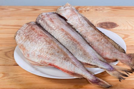 Cooled carcasses of the red cod without heads, cleared from fish scales on white dish on a wooden rustic table, side view in selective focus