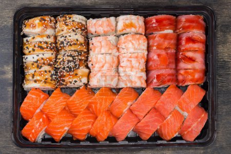 Set der verschiedenen Sushi in offener schwarzer Plastikverpackung zum Mitnehmen auf schwarzer Oberfläche, Nahaufnahme von oben