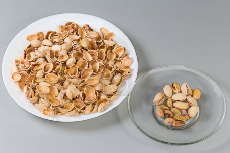 Partly peeled roasted salted pistachio nuts on the glass saucer against the empty shells on a big dish on a gray background