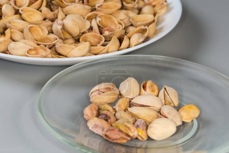 Partly peeled roasted salted pistachio nuts on the glass saucer against the empty shells on a big dish on a gray background, fragment side view in selective focus