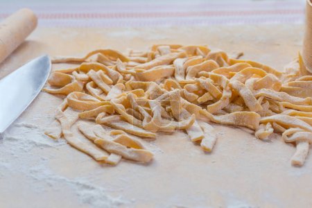 Heap of the raw freshly prepared homemade egg noodles in the shape of the long strips on a wooden board sprinkled with flour, side view in selective focus