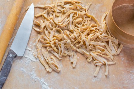 Heap of the raw freshly prepared homemade egg noodles in the shape of the long strips on a wooden board sprinkled with flour, top view
