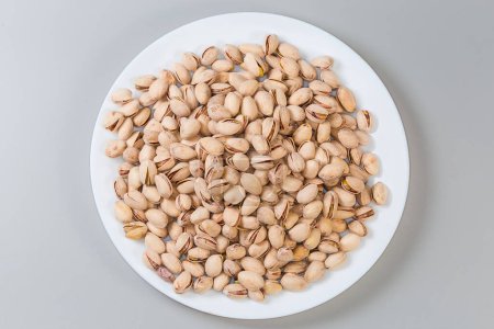 Heap of the roasted salted pistachio nuts with partly naturally open shells on a big white dish on a gray background, top view