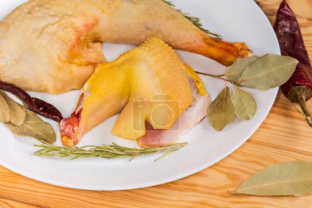 Rohe Hahnenkeule und Hahnenschulter, die im Freilandbetrieb angebaut werden, mit gelber Schale zwischen den verschiedenen trockenen Gewürzen auf einem rustikalen Tisch, Fragment in Großaufnahme