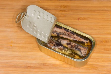 Canned sardines in cooking oil in small partly opened tin can with ring-pull tab lid on a wooden surface
