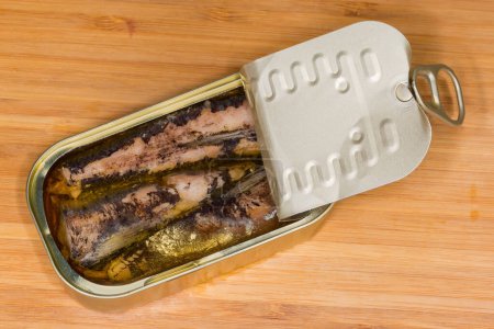Canned sardines in cooking oil in small partly opened tin can with ring-pull tab lid on a wooden surface, top view