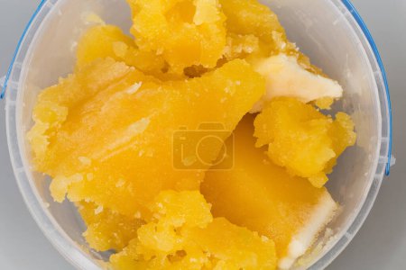 Pieces of crystallized white honey in open round plastic container on a gray background, top view of fragment close-up