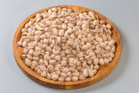 Heap of the roasted salted pistachio nuts with partly naturally open shells on a big wooden dish on a gray background