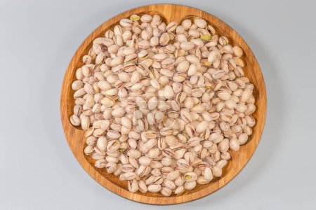 Heap of the roasted salted pistachio nuts with partly open shells on a big wooden dish on a gray background, top view 