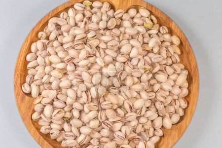 Heap of the roasted salted pistachio nuts with partly open shells on a big wooden dish on a gray background, top view of fragment close-up