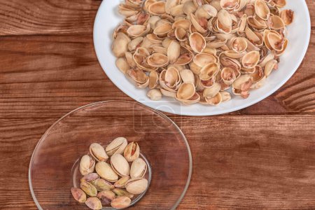 Partly peeled roasted salted pistachio nuts on the glass saucer and separately empty shells on a big dish on a rustic table