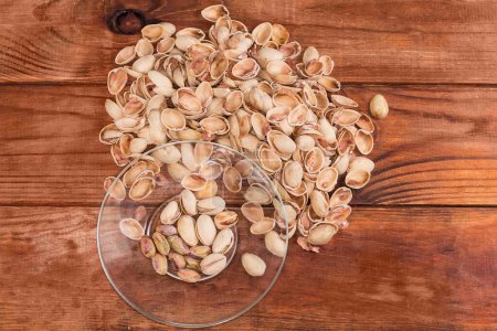 Partly peeled roasted salted pistachio nuts on the transparent glass saucer against the empty shells on a rustic table, top view 