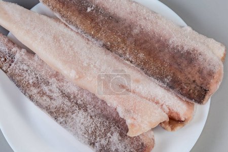 Raw frozen fillets of hake in the ice glaze covered with hoarfrost on plate on a gray background, top view of fragment close-up