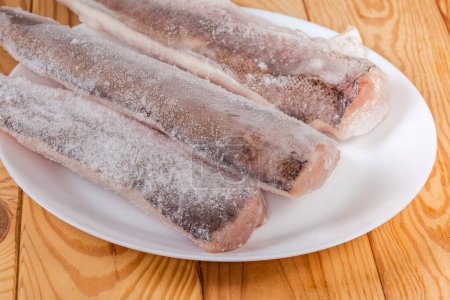 Roh gefrorene Seehechtfilets in der Eisglasur mit Raureif auf weißem Teller auf dem rustikalen Tisch, Nahaufnahme in selektivem Fokus
