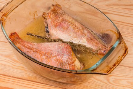 Two baked redfishes without heads, also known as ocean perch in the old glass baking dish on a rustic table, fragment close-up
