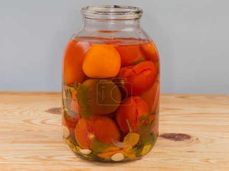 Tomates en conserve de différentes variétés avec des épices et des légumes dans le grand pot en verre ouvert sur une table rustique, vue de côté sur un fond gris