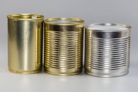 Diferentes latas vacías abiertas de debajo de una comida enlatada, con varias cubiertas blancas y amarillas sobre un fondo gris, vista lateral