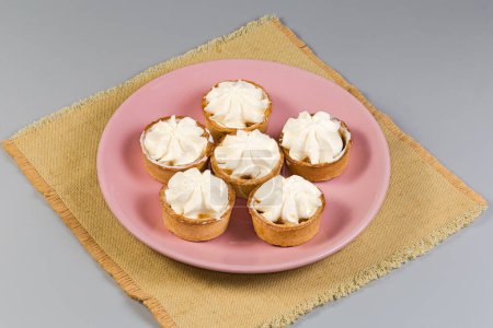 Small tarts, so-called tartlets made with baked short crust pastry molds, caramelized pear filling and custard on top on pink dish on a napkin on a gray background