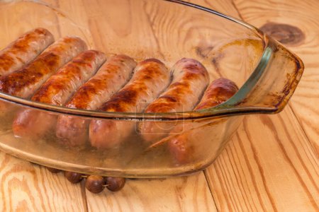 Baked thick pork sausages in natural casing on the old glass casserole pan on a rustic table, fragment close-up in selective focus