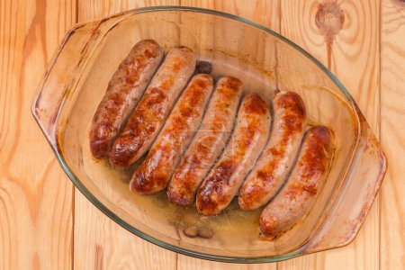 Baked thick pork sausages in natural casing on the old glass casserole pan on a rustic table, top view